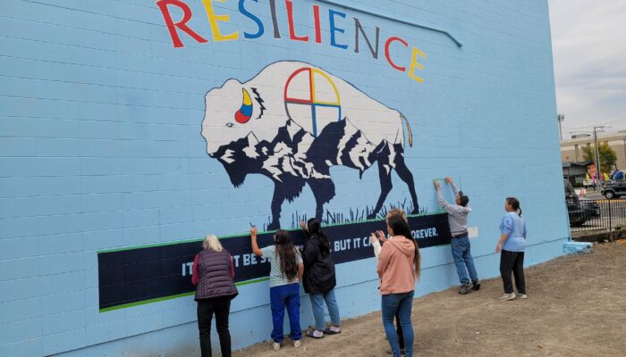 The word resilience is written in color overhead a 40 foot black and white bison mural. Paintets work on the finishing touches of the bottom of the mural that says, it might be stormy now, but it wont rain forever.